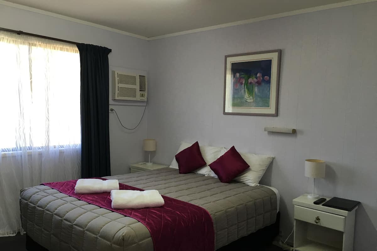 A hotel room with a bed, a nightstand, a lamp, a painting, and an air conditioning unit.