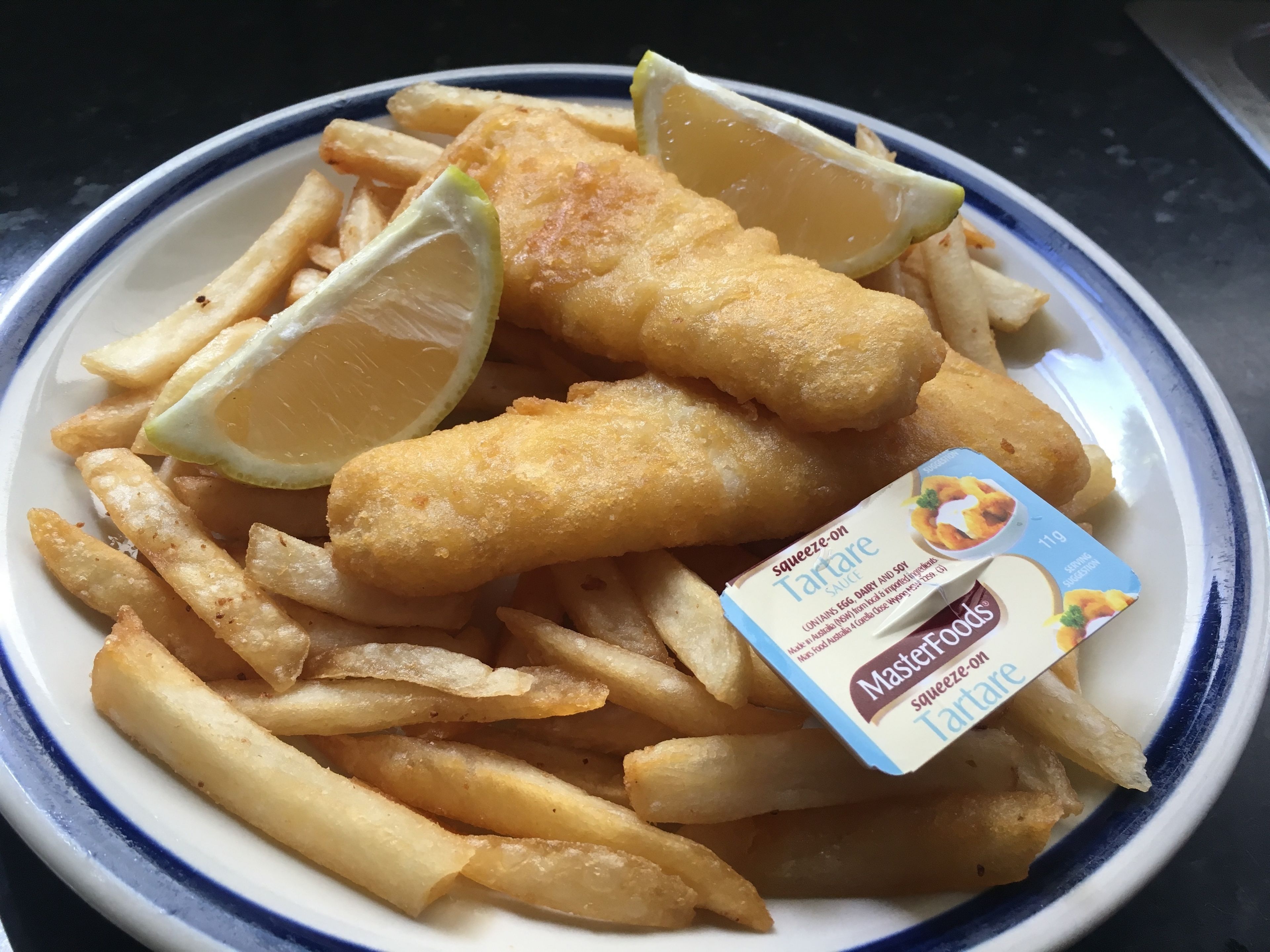 A plate of fish and chips with lemon wedges and a squeeze-on tartare sauce packet.