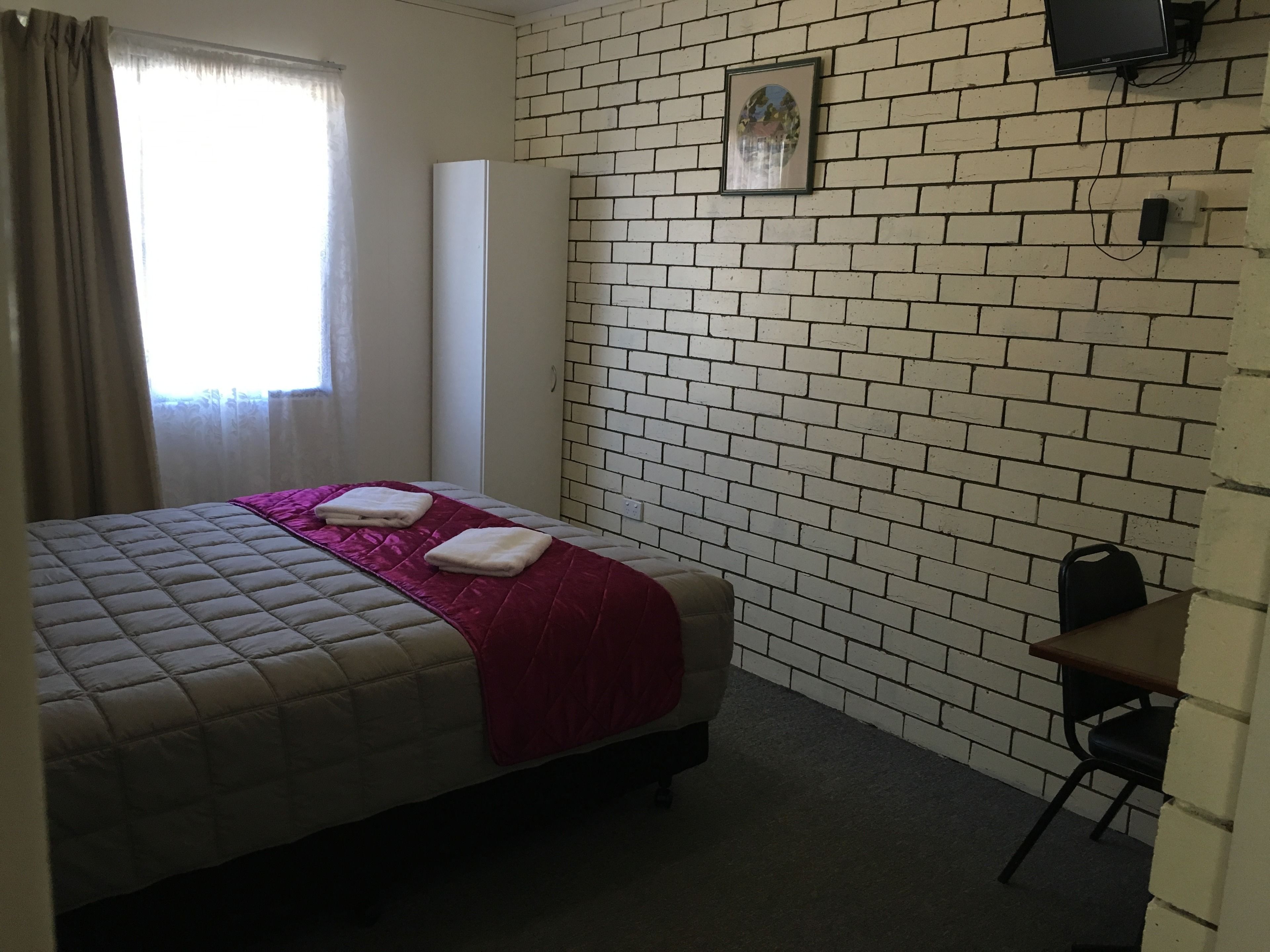 A single bed with a quilted headboard, a red throw blanket, and two folded towels. A small desk with a chair is against the brick-patterned wall.