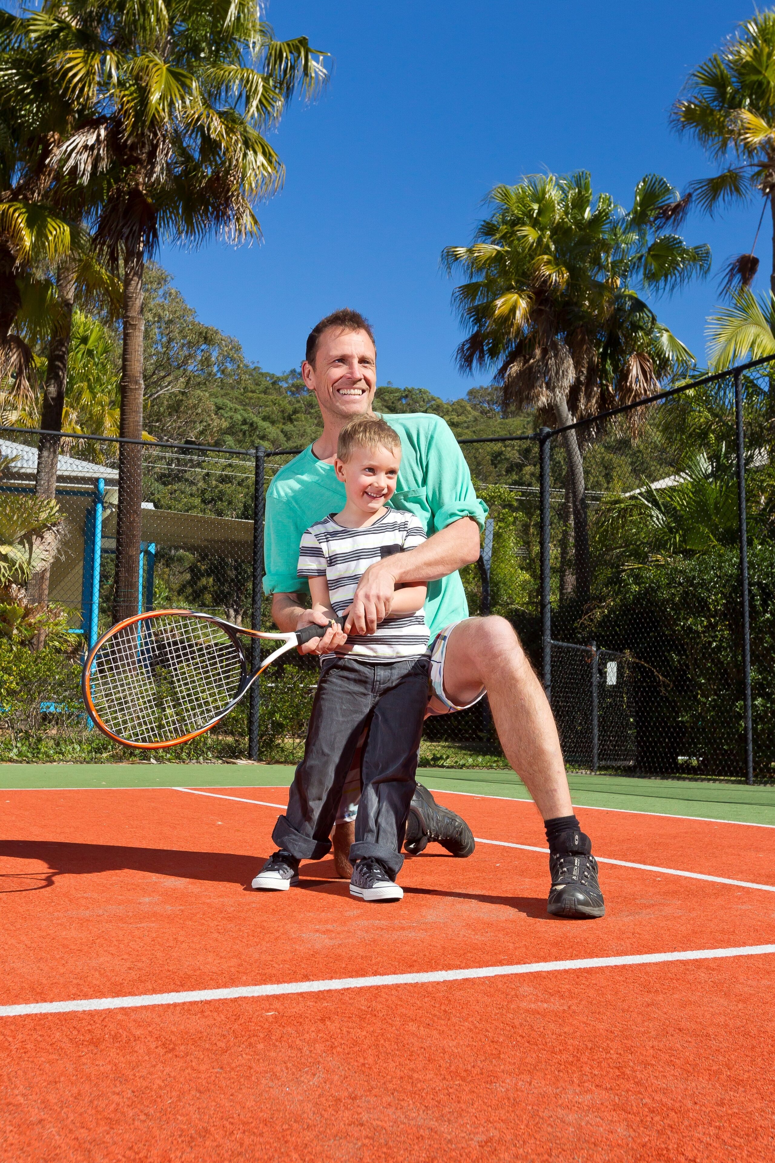 En tennisbana med ett nät, ett barn som håller i ett tennisracket och en man som hukar sig bredvid barnet.