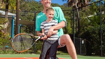 Un terrain de tennis avec un filet, un enfant tenant une raquette de tennis et un homme accroupi à côté de lui.