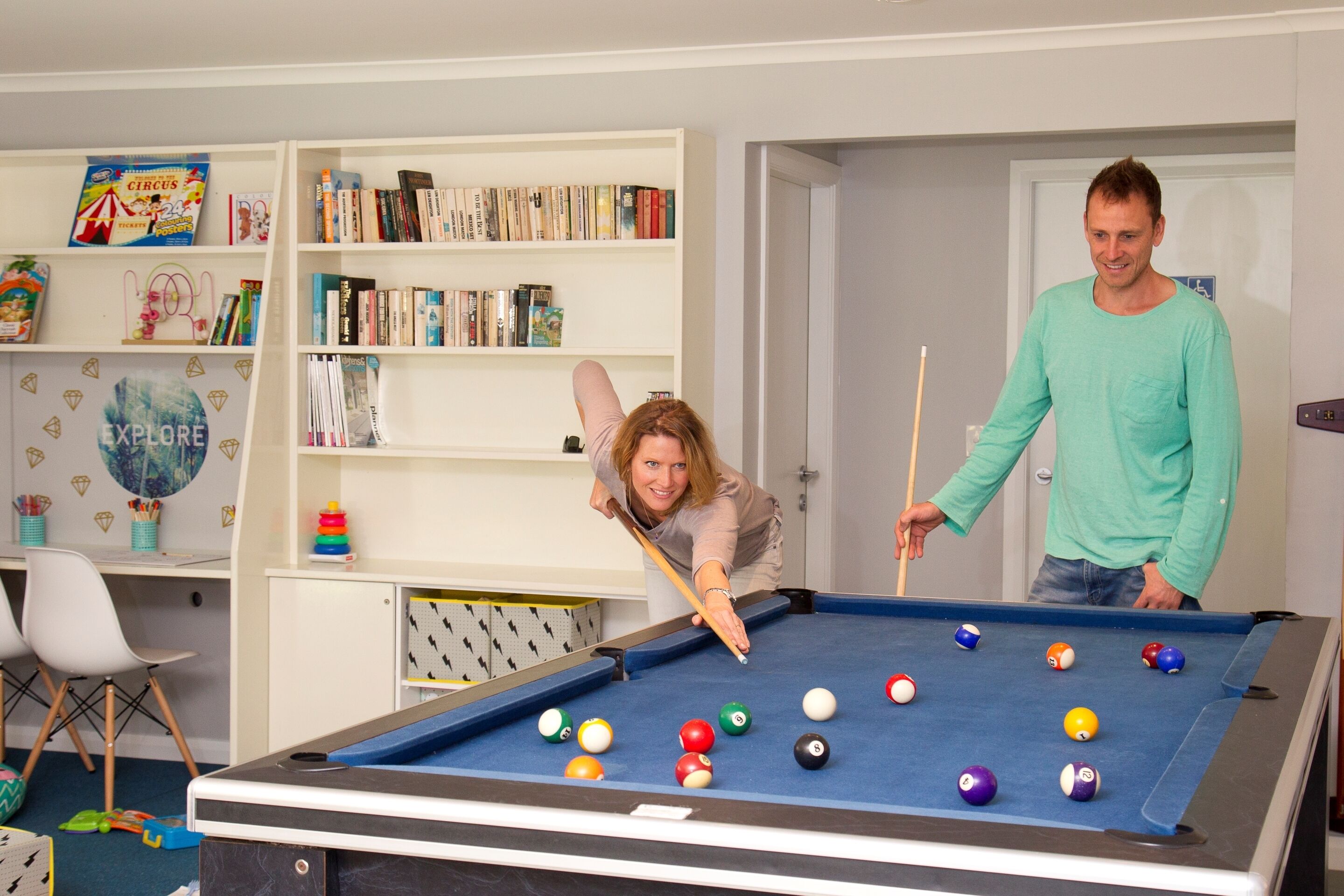 A pool table with various colored balls, a bookshelf with books and a circus-themed book, and a door in the background.