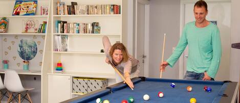 A pool table with various colored balls, a bookshelf with books and a circus-themed book, and a door in the background.