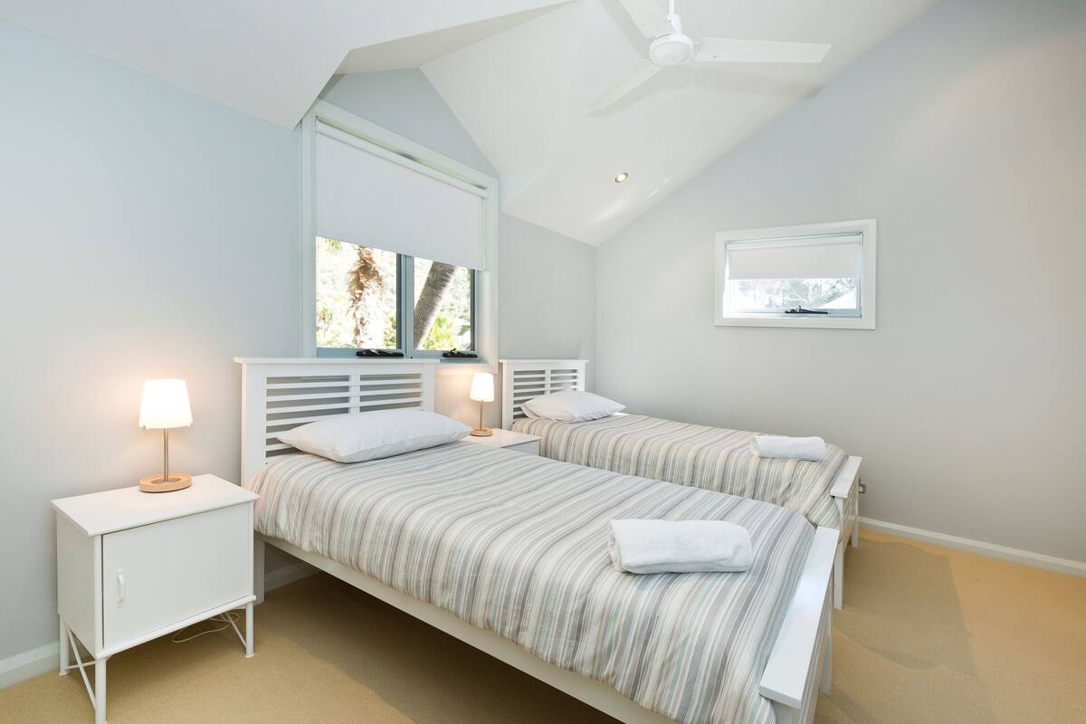 A bedroom with two beds, a ceiling fan, a window, and a nightstand with a lamp.
