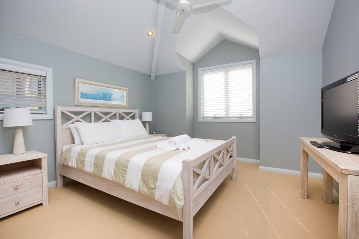 A bedroom with a bed, a TV, a desk, a ceiling fan, a window, and a nightstand.
