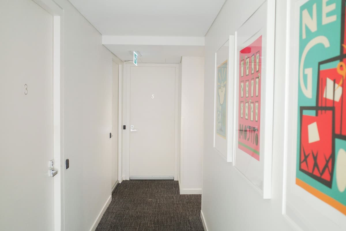 Un couloir aux murs blancs, doté d’un sol recouvert de moquette et de portes numérotées. Des œuvres d’art encadrées ornent les murs.