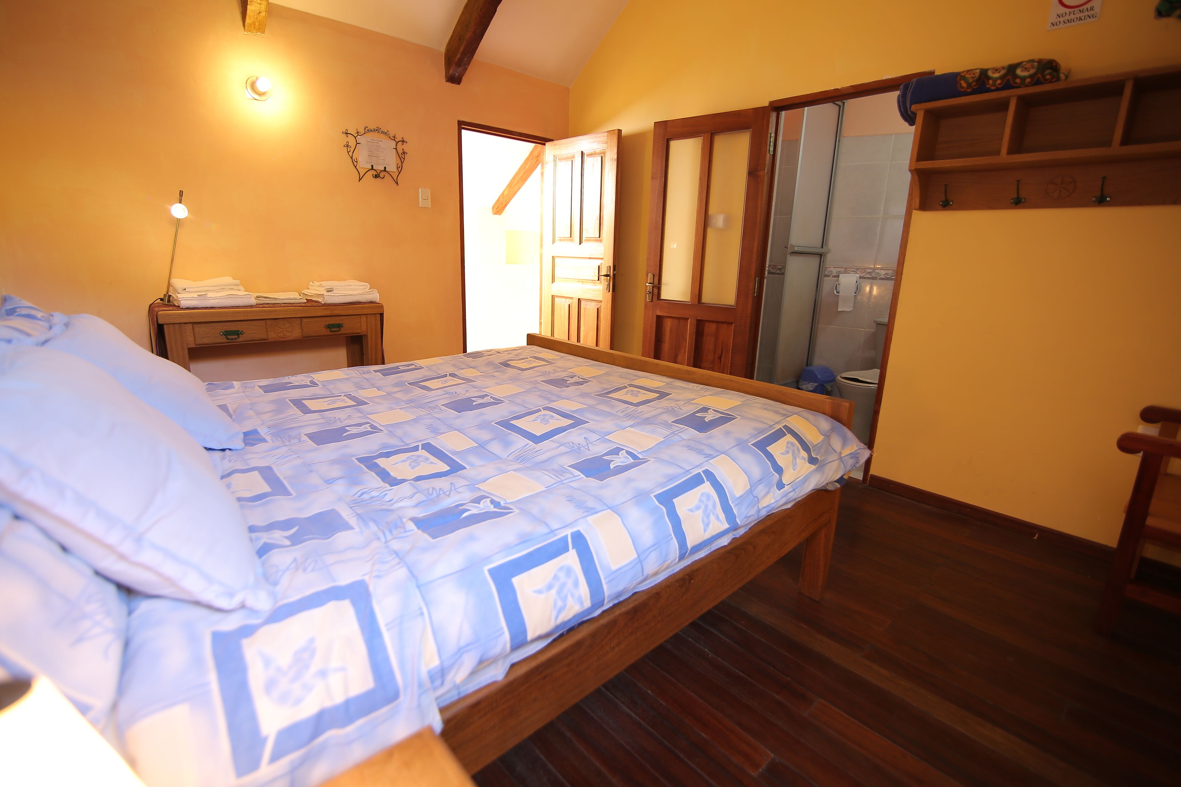 Une chambre à coucher comprenant un lit, une tête de lit en bois, une table de chevet, une lampe fixée au mur et une porte donnant accès à une salle de bain.