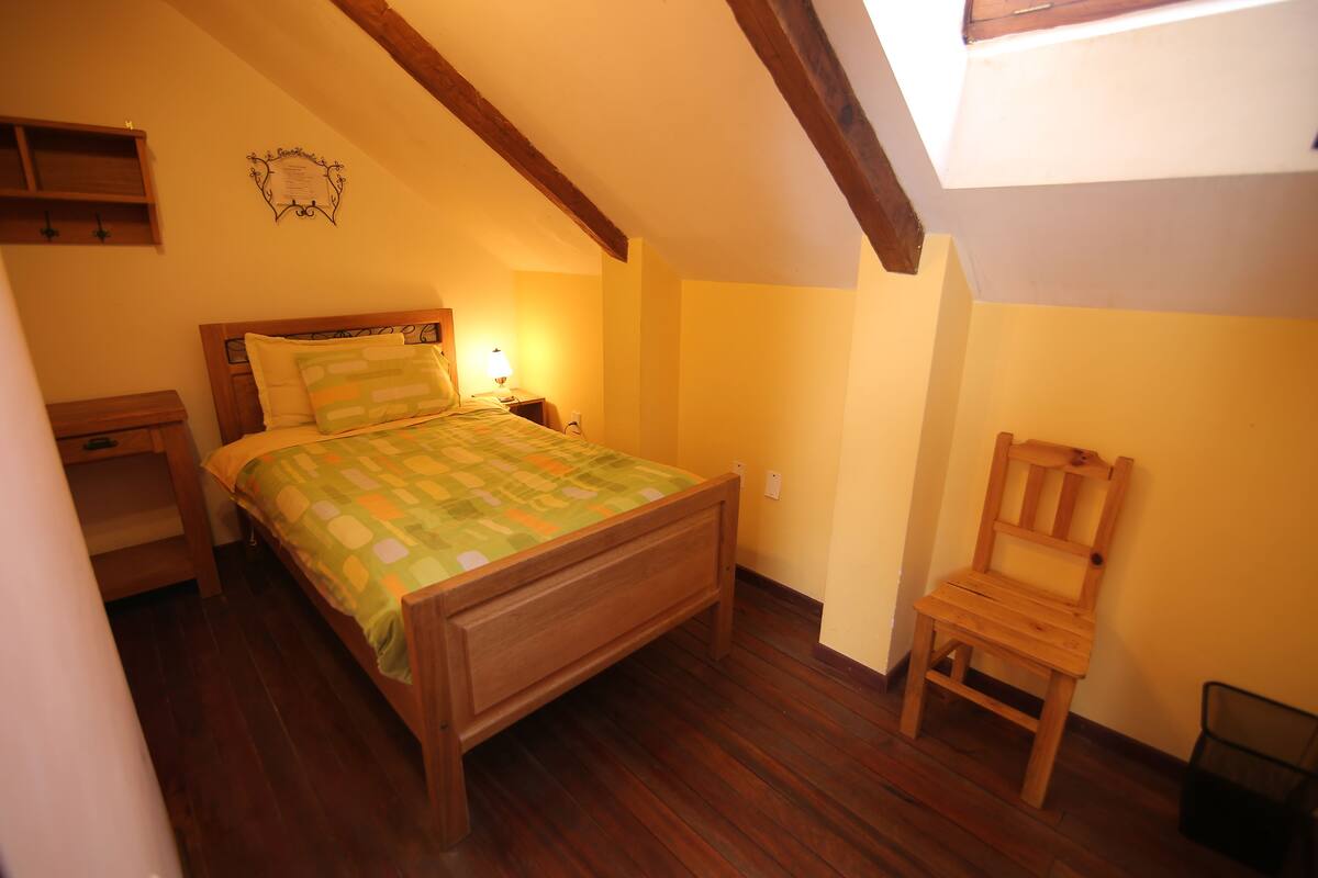 A bedroom with a wooden bed, a nightstand, a chair, and a sloped ceiling with exposed beams.