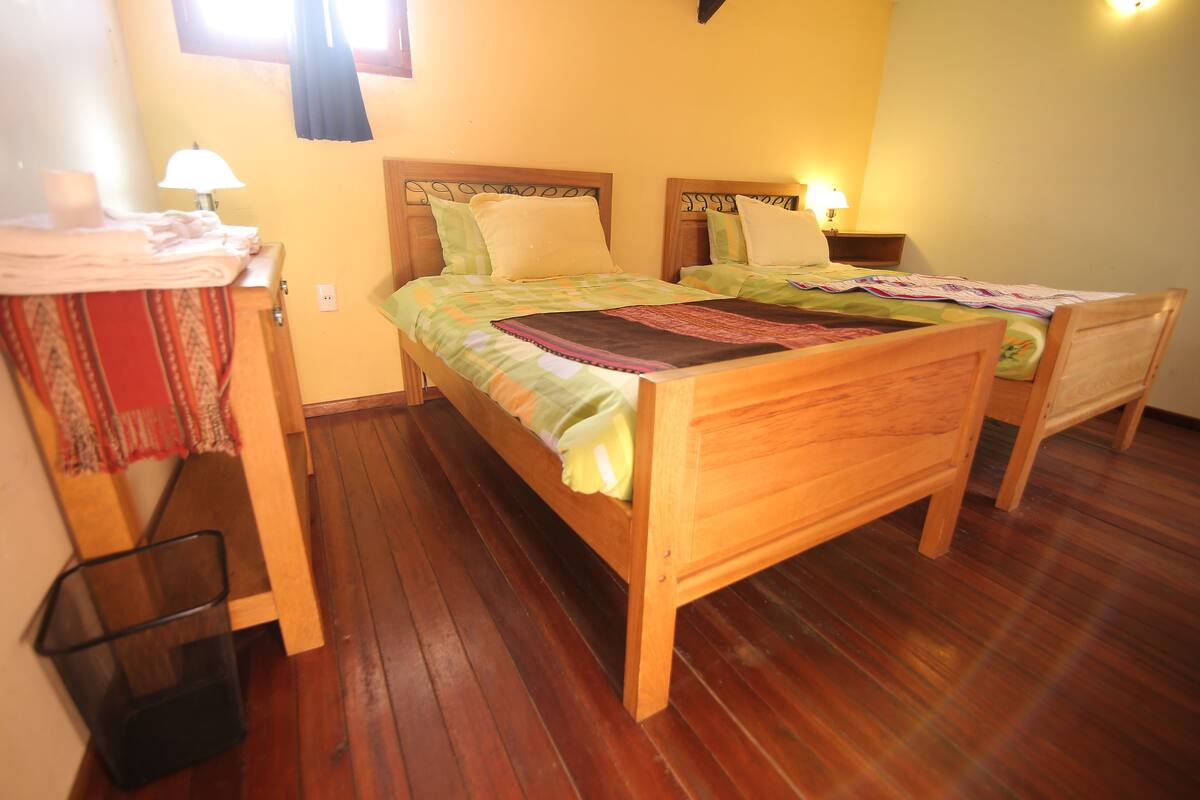 A room with two wooden beds, each with a headboard and a bedside table with a lamp. There is a small trash bin and a wooden chair.