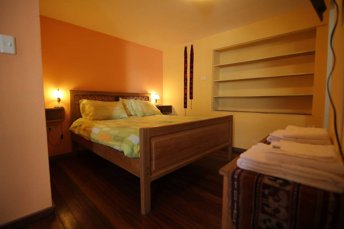 A bedroom with a wooden bed, two bedside lamps, a wall-mounted shelf, and a window with blinds.