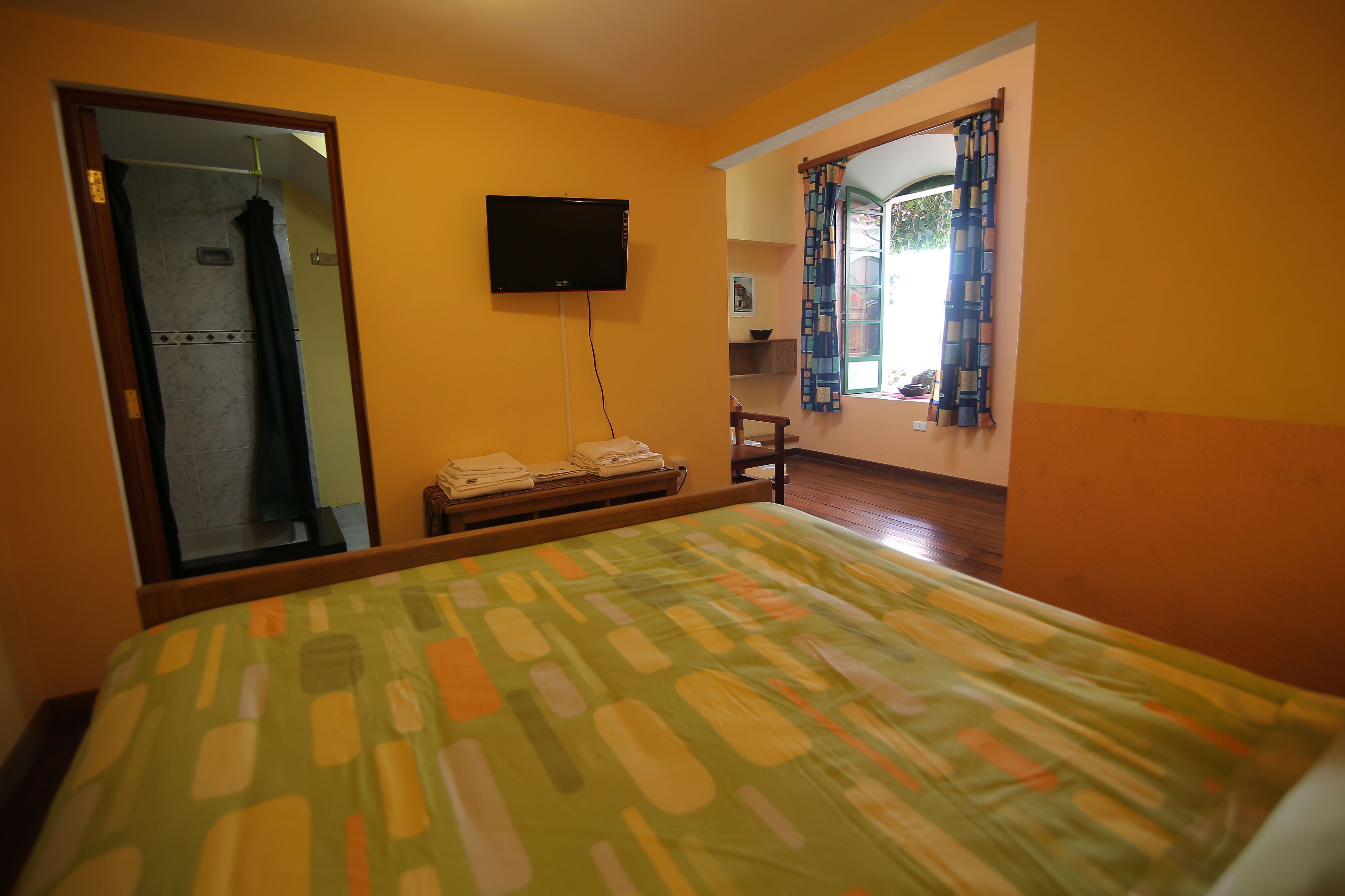 A hotel room with a bed, a television mounted on the wall, a window with curtains, and a small seating area.