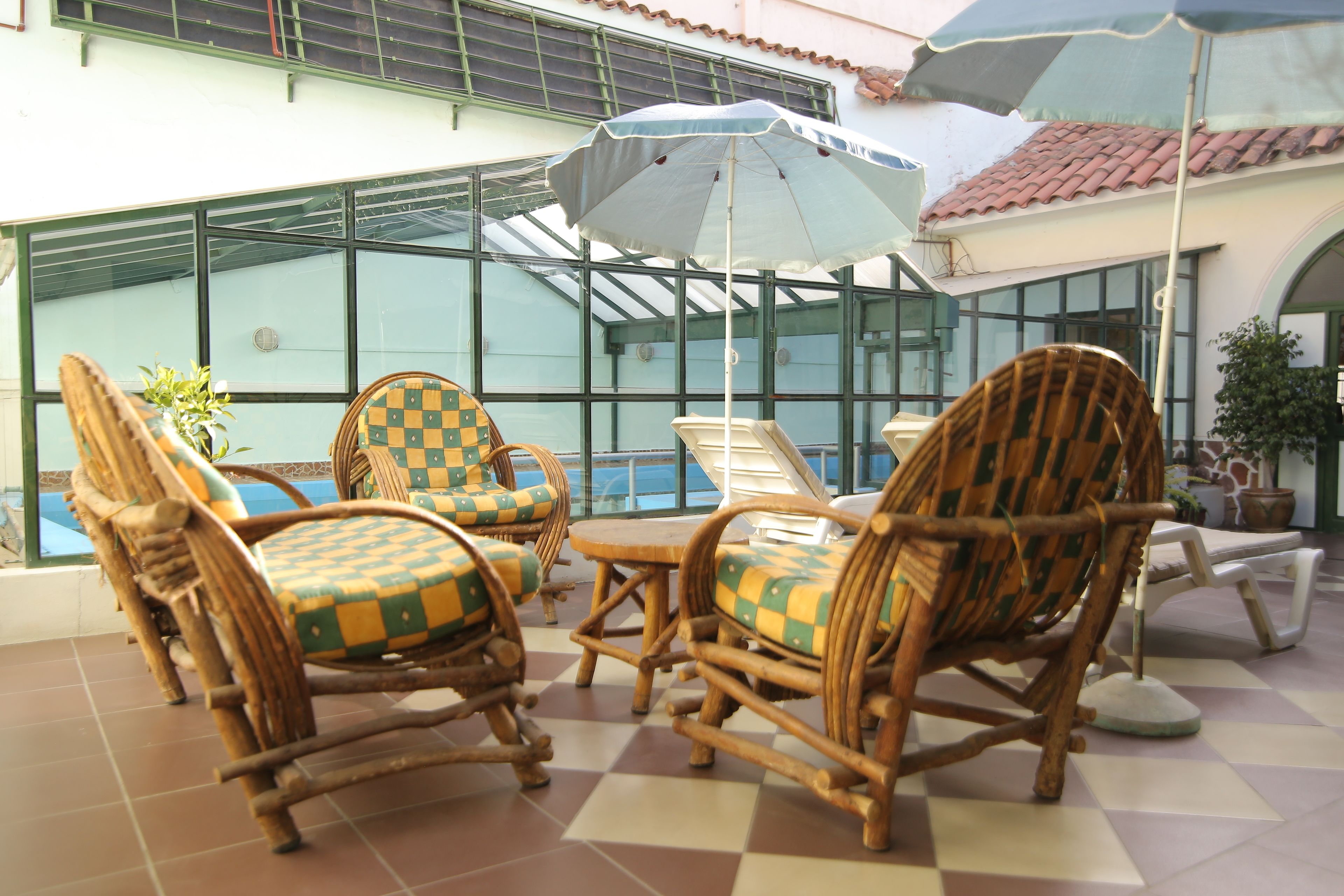 Des chaises en osier avec des coussins à carreaux, une table ronde et des parasols dans un espace extérieur abrité, avec vue sur la piscine.