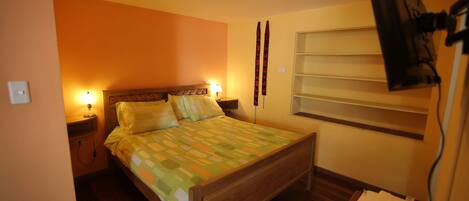 A single-bed room with a wooden headboard, a bedside table with a lamp, and a window with blinds.