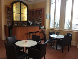Un coin bar agrémenté d’une fenêtre à vitraux, d’étagères en bois où sont exposées des bouteilles, et de deux tables rondes avec des chaises.