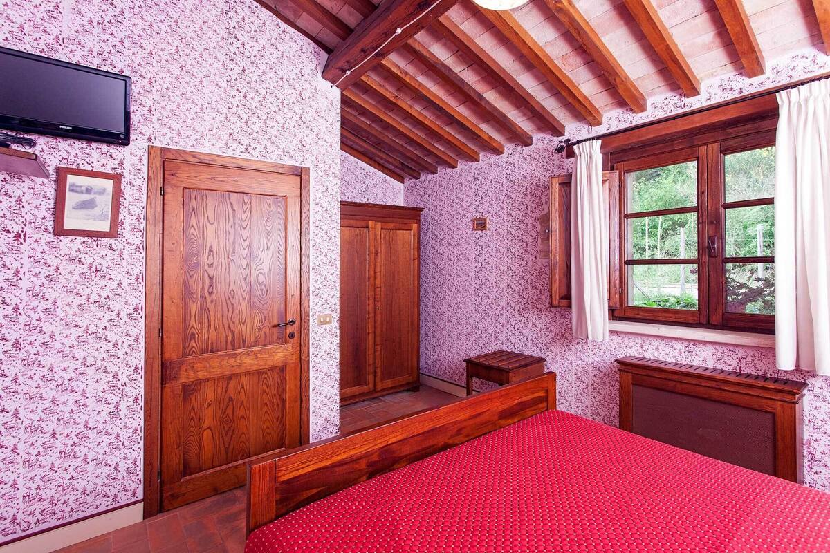 Un dormitorio con techo de madera, una cama roja, un armario de madera y una ventana con cortinas.