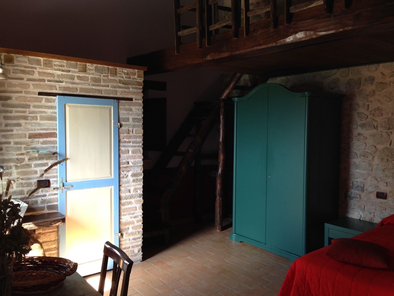 A room with a stone wall, a wooden door, a green wardrobe, a wooden chair, and a red bed.