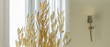 A decorative golden plant in a stone pot on a glass table near a window with white frames.