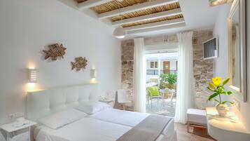 A modern hotel room with a large bed, a stone wall, a balcony with outdoor seating, and a view of the sea.
