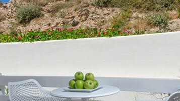 Una mesa blanca al aire libre con un plato de manzanas verdes, rodeada de sillas blancas, con un paisaje de colinas rocosas y cielo azul despejado de fondo.