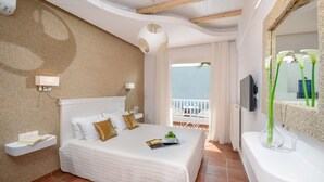 A hotel room with a bed, a bedside table, a TV, a balcony with a white railing, and a view of the sea. - Adriani Hotel (Naxos)