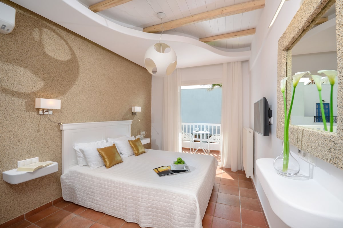 Une chambre d’hôtel avec un lit, une table de chevet, une télévision, un balcon doté d’une balustrade blanche et une vue sur la mer.