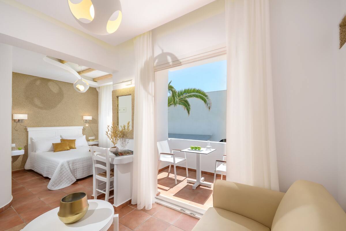 Une chambre d’hôtel avec un lit, une petite table avec des chaises, un balcon donnant sur la mer et une fenêtre avec des rideaux.