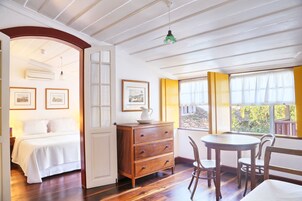 Una habitación con cama, cómoda de madera, mesa de comedor y sillas. Se observan cuadros en la pared y una ventana con cortinas.