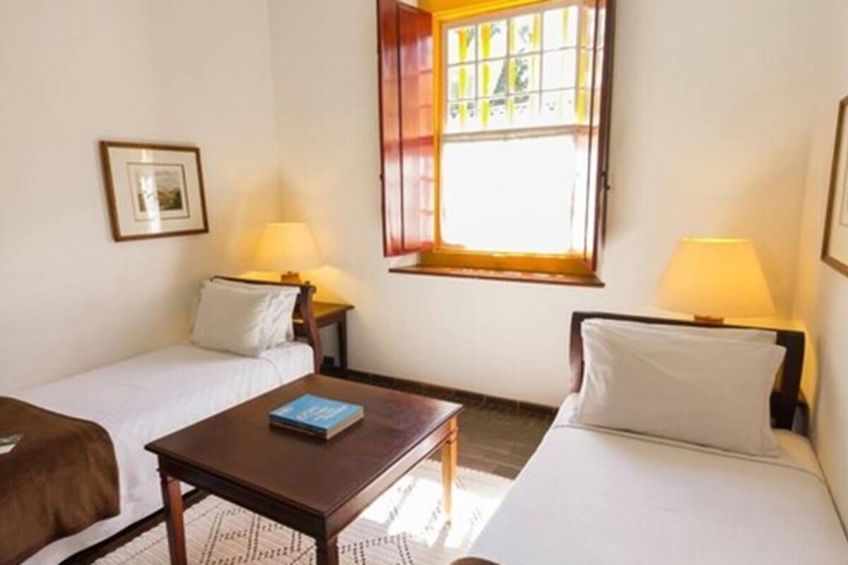 A room with two single beds, a small table, two bedside lamps, and a window with curtains.