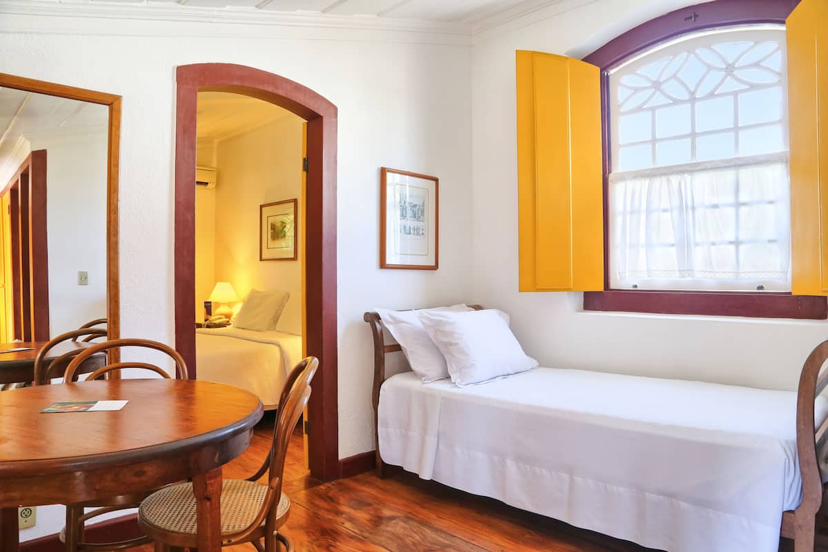 A room with a single bed, a dining table with chairs, a mirror, and a window with yellow shutters.