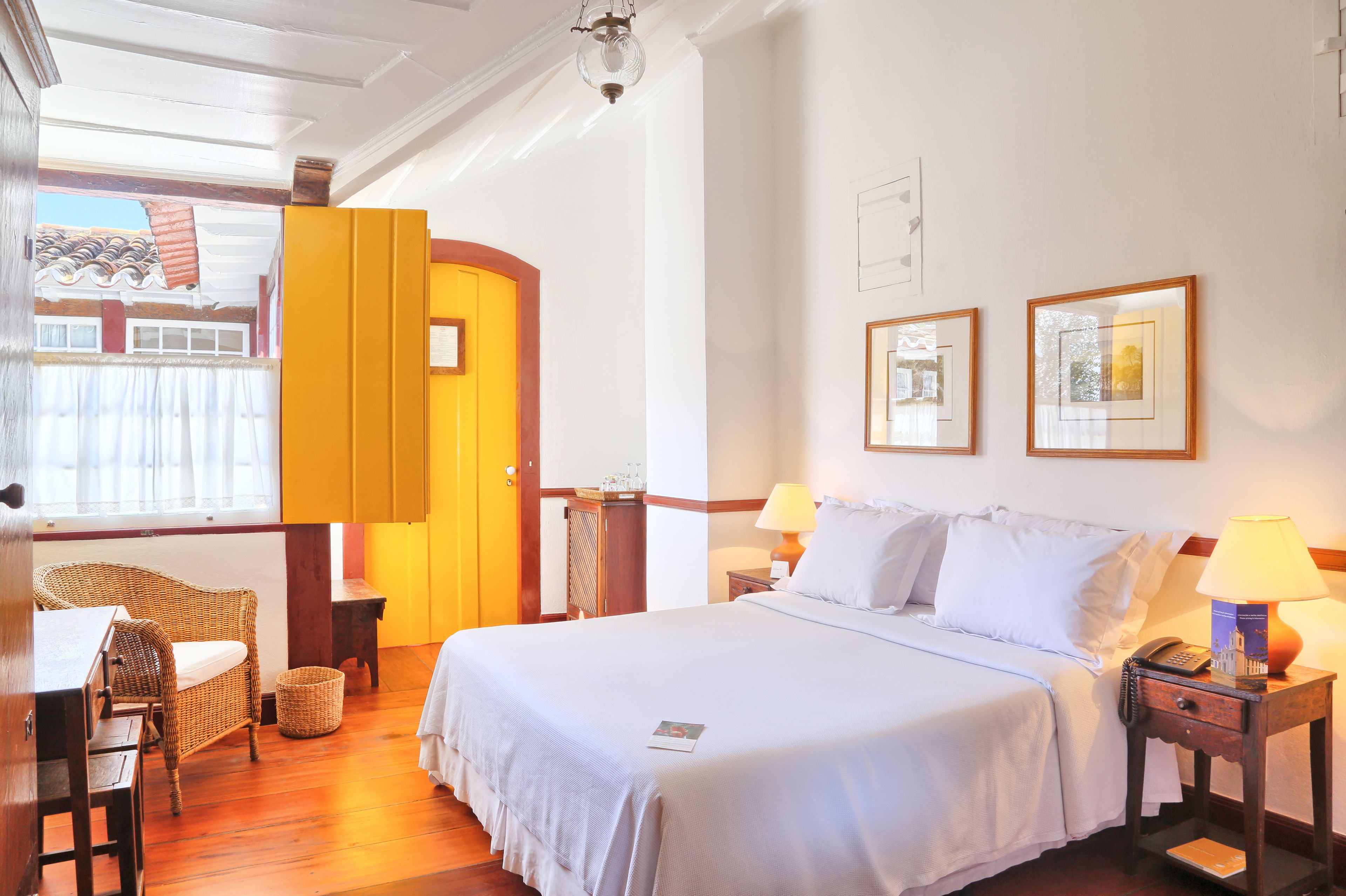 A hotel room with a large bed, two bedside tables with lamps, a wicker chair, a small table, and a window with curtains.