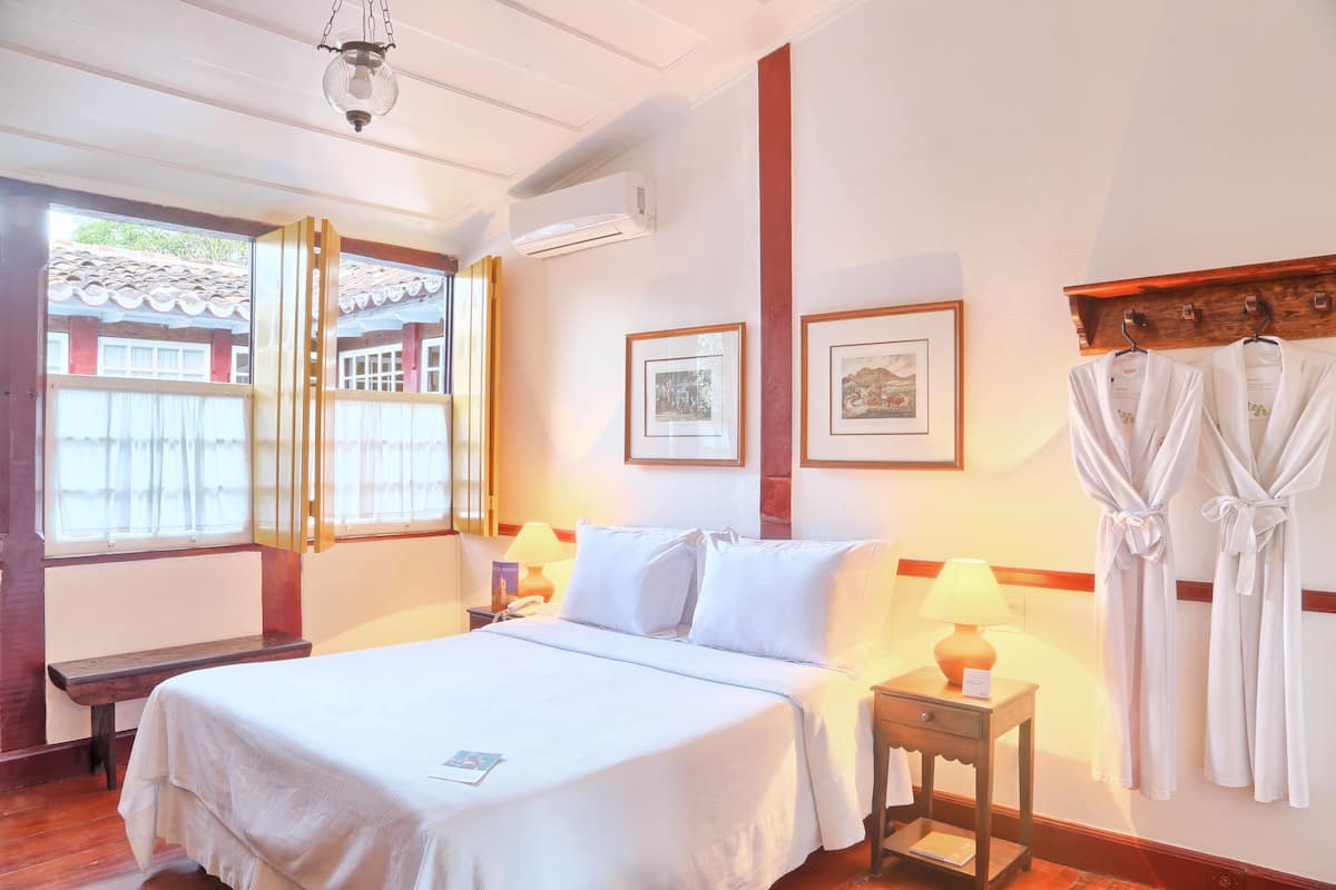 A hotel room with a bed, bedside table, lamp, robe hooks, and a window with curtains.