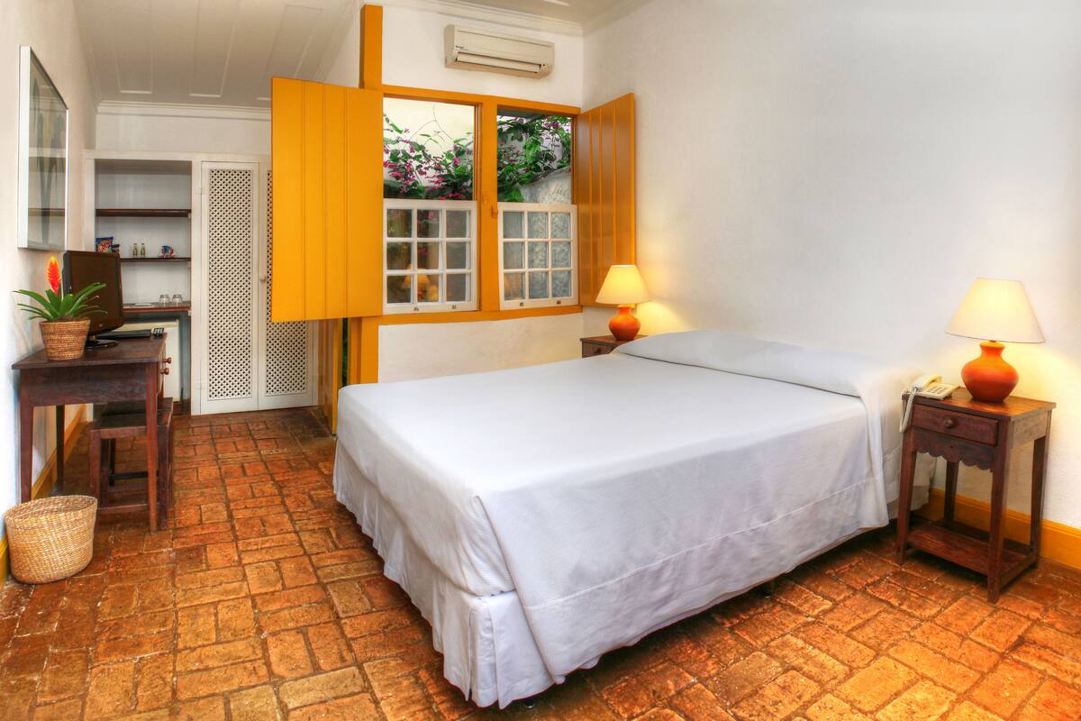 A bedroom with a bed, two bedside tables with lamps, a desk with a chair, a potted plant, and a window with open shutters.