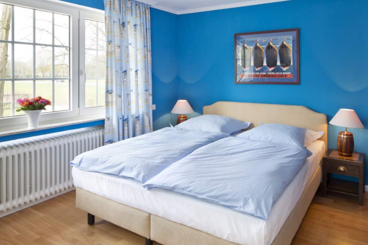 Un dormitorio con una cama grande, dos mesitas de noche, una ventana con cortinas, un radiador y un cartel en la pared.