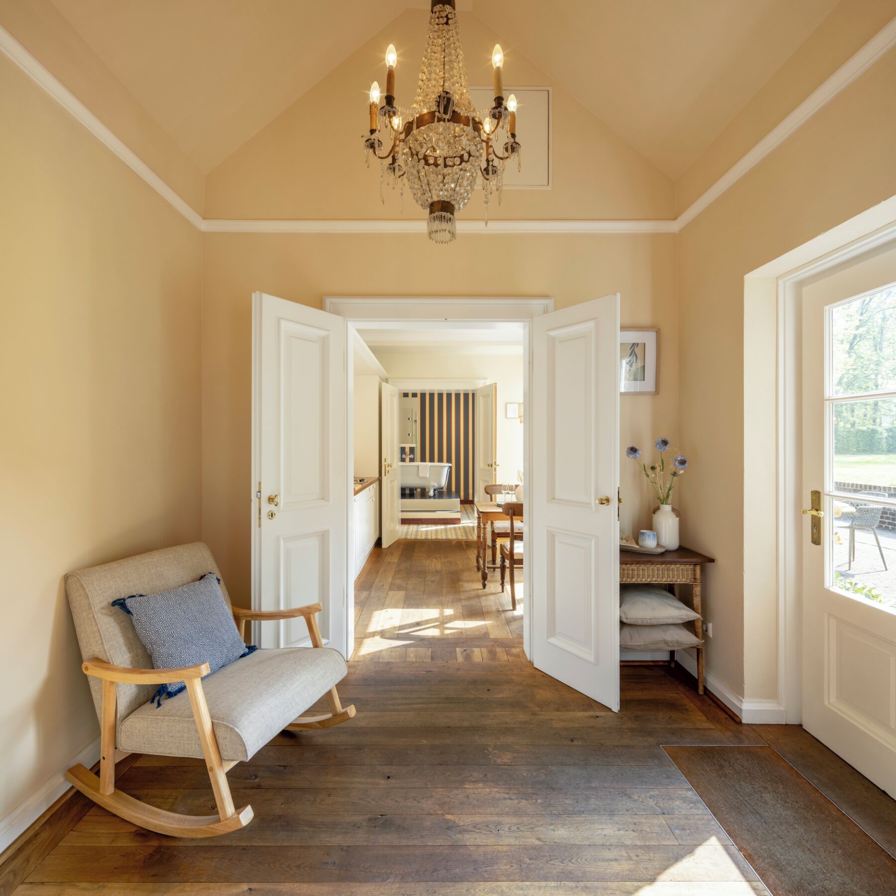 A hallway with a wooden rocking chair, a chandelier, and a view into a dining area.