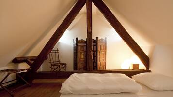 Quarto com teto inclinado, estrutura de cama em madeira, roupa de cama branca, uma cadeira e um biombo decorativo.