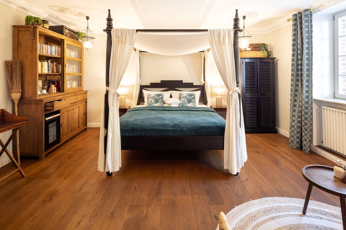 Una cama con dosel y cuatro columnas con un edredón verde oscuro, estanterías de madera, una mesita y una cortina estampada.