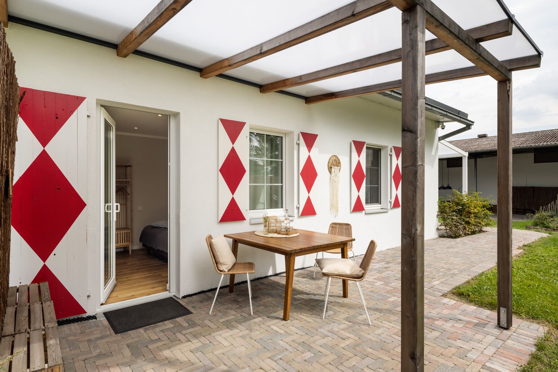 A covered patio with a wooden table and chairs, a white wall with red diamond patterns, and a door leading to an interior room.