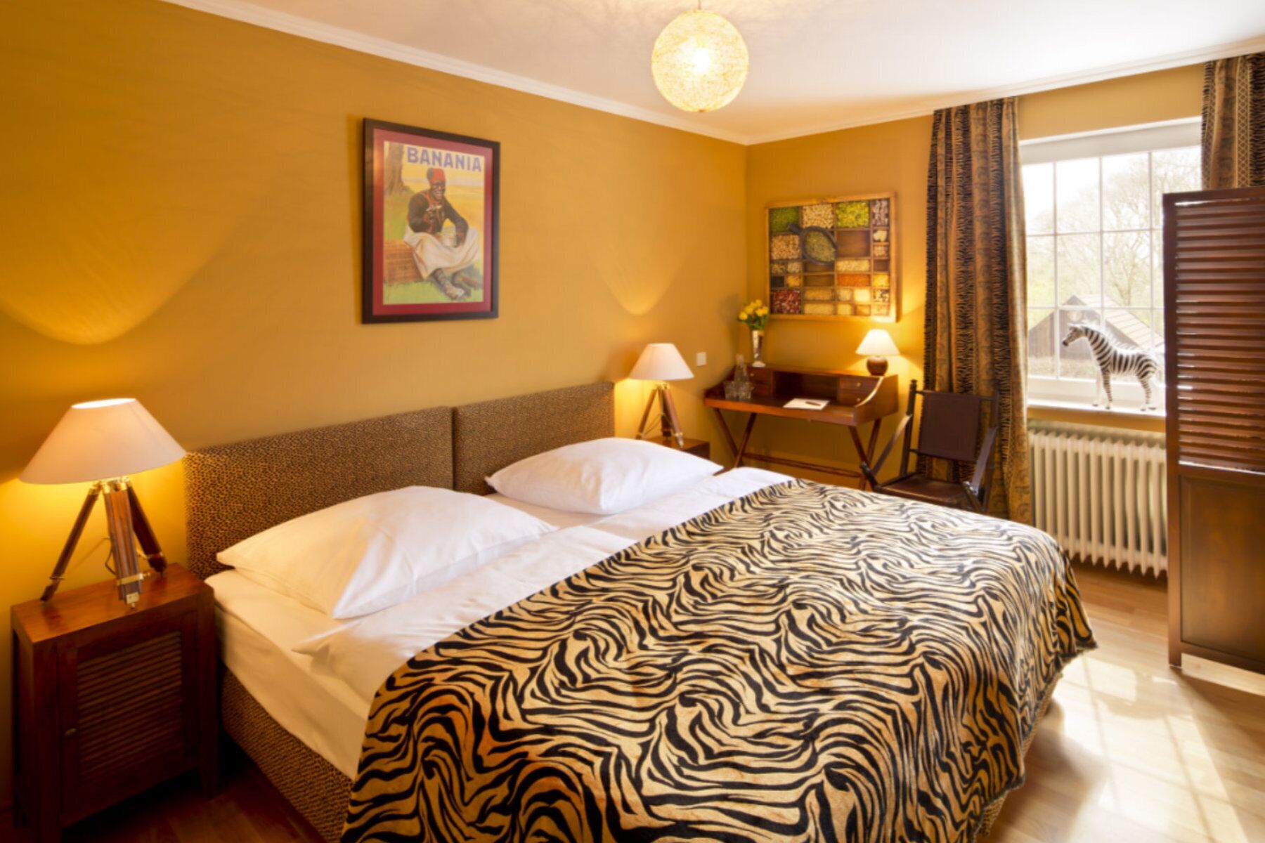 A hotel room with a bed, two bedside lamps, a zebra-patterned bedspread, a framed picture on the wall, a desk with a lamp, and a window with a zebra outside.