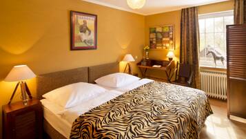 A hotel room with a bed, two bedside lamps, a zebra-patterned bedspread, a framed picture on the wall, a desk with a lamp, and a window with a zebra outside.