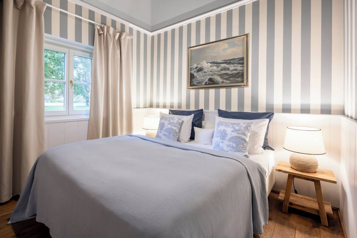 Un dormitorio con una cama grande, papel tapiz a rayas, un cuadro de un paisaje marino y una ventana con cortinas.
