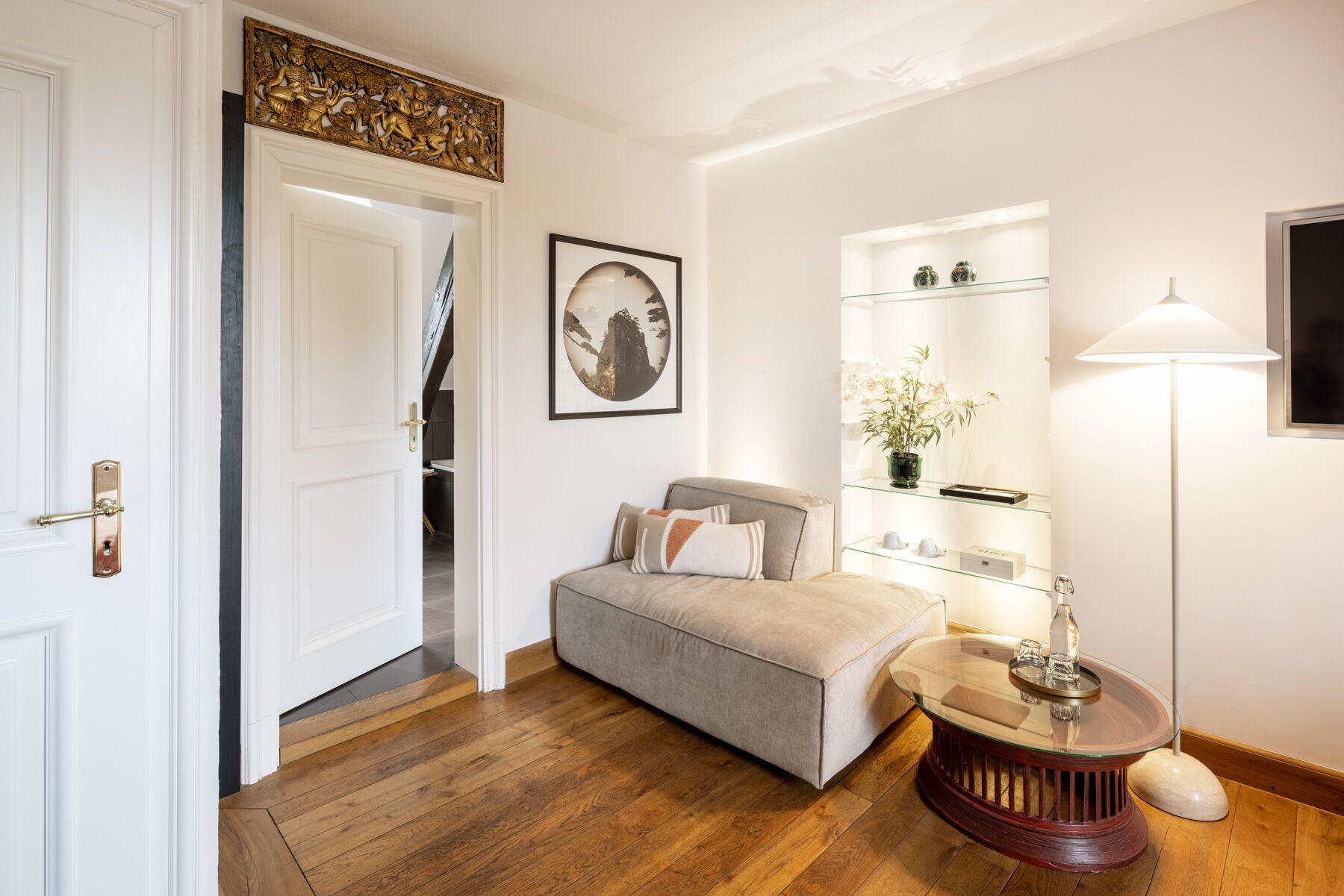 A room with a wooden floor, a beige sofa, a glass coffee table, a lamp, and a shelf with decorative items.
