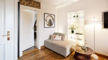 A room with a wooden floor, a beige sofa, a glass coffee table, a lamp, and a shelf with decorative items.