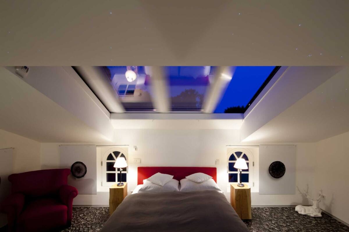 Una habitación de hotel con tragaluz, cabecera roja, dos mesitas de noche con lámparas, un sillón rojo y una alfombra estampada.