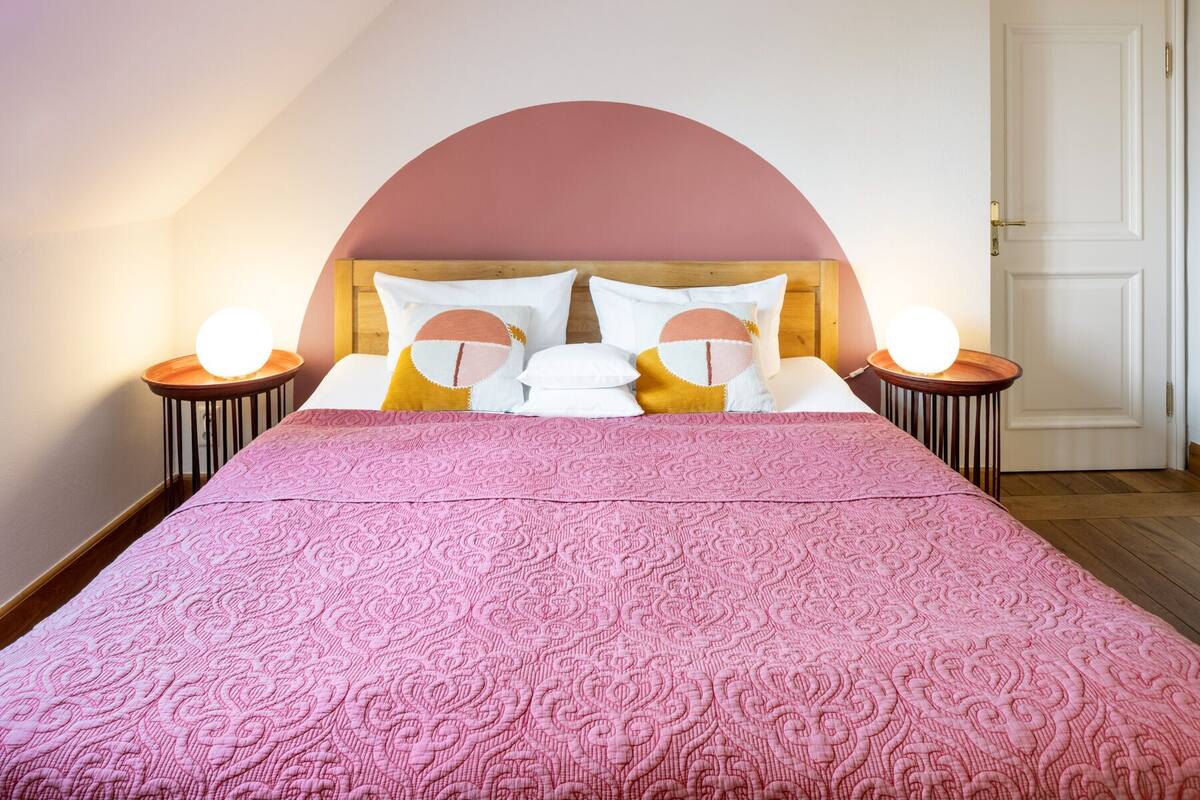 Una cama bien hecha con un edredón rosa, dos mesitas de noche con lámparas redondas y una puerta blanca al fondo.