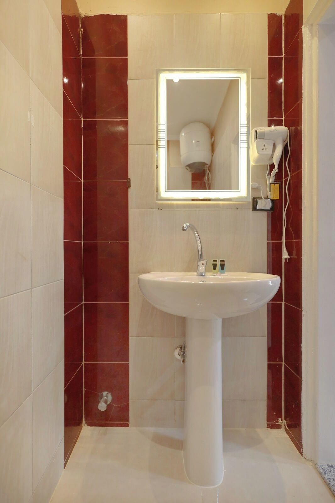 Un baño con lavamanos pedestal blanco, un espejo con luces y un secador de pelo en la pared.