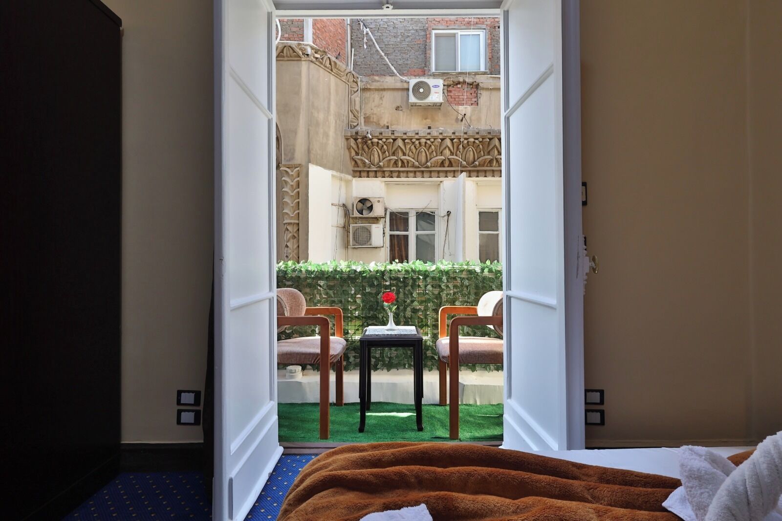 A hotel room with a bed, a brown blanket, and a view of an outdoor seating area with two chairs and a small table.