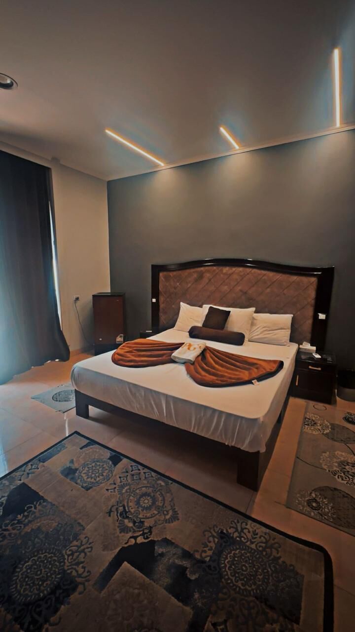 A modern hotel room with a large bed, a bedside table, a nightstand, and a patterned rug.