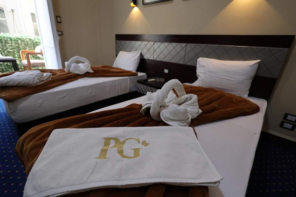 A hotel room with two beds, each with a brown blanket and white pillows. There is a towel with 'PG' on it on the floor.