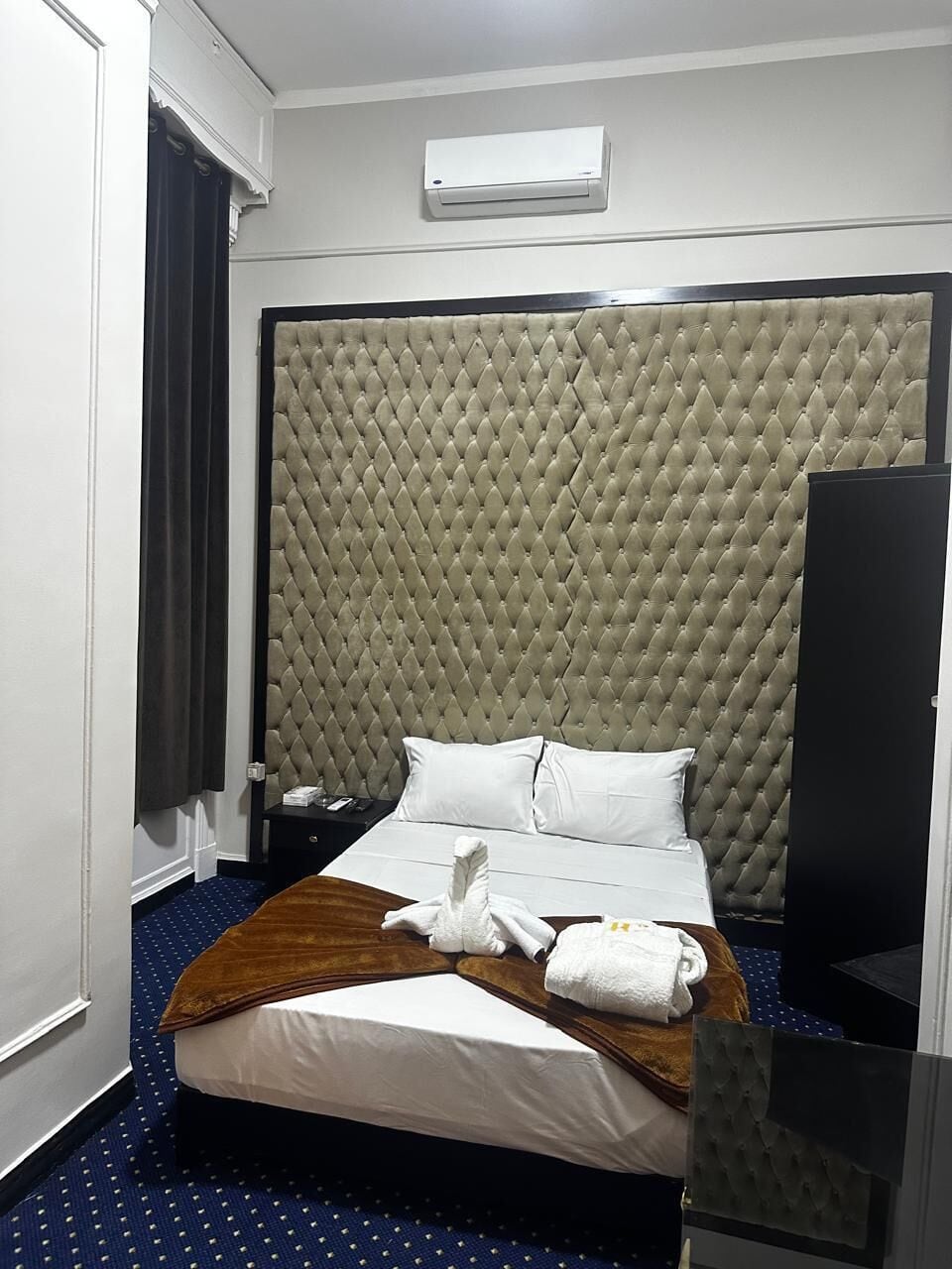A hotel room with a large bed, a headboard, a bedside table, an air conditioning unit, and a window with curtains.