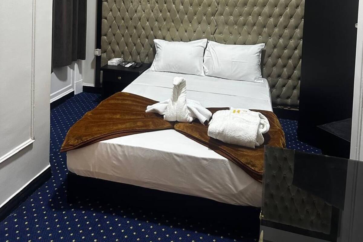 A hotel room with a large bed, a headboard, a bedside table, an air conditioning unit, and a window with curtains.